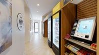 This single-floor commercial space is designed with functionality and convenience in mind. A bright, clean corridor with light-toned tile flooring runs the length of the space, creating an open and welcoming flow toward a glass-paneled exit door at the far end. Recessed lighting overhead ensures the area is well-lit throughout.

The standout feature of the space is a thoughtfully arranged refreshment station positioned along one side of the hallway. Warm wood-paneled cabinetry and shelving provide a structured backdrop for two large glass-door refrigerators stocked with beverages, clearly branded under signage labeled "hydrate" and "nourish" in bold yellow and green tones. Adjacent shelving units display a variety of packaged snacks and convenience items, accompanied by a self-service kiosk for easy transactions.

The overall aesthetic balances practicality with a modern, clean design. The contrast between the warm wood tones and the bright white walls and flooring gives the space a fresh, approachable ambiance. Whether serving hotel guests, office workers, or retail customers, this space is purpose-built to provide quick and easy access to refreshments and essentials in a tidy, organized environment.