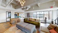 Located at 707 S Fourth St in Champaign, Illinois, this single-floor amenity space is designed with a contemporary industrial aesthetic, showcasing exposed concrete ceilings, visible ductwork, and an open layout that promotes a lively, social atmosphere.

The game room is a standout feature, centered around a full-size pool table set atop striking geometric patterned tile flooring. Large floor-to-ceiling windows flood the area with natural light and offer views of the surrounding Champaign cityscape. A foosball table and additional seating areas complete the recreational offerings.

Adjacent to the game room, the lounge area provides a more relaxed setting, anchored by a sleek built-in fireplace framed by a dark surround and flanked by open wood shelving. A wall-mounted TV sits above the hearth, while plush sofas, ottomans, and curated artwork contribute to a warm and inviting ambiance. Artistic pendant chandeliers and mixed hardwood flooring add warmth and character throughout.

The communal dining and kitchenette area features a long wooden dining table with seating for a group, pendant lighting overhead, and a wet bar kitchenette equipped with countertop appliances. Bold wallcoverings and expressive wall art give the space a distinct personality. Together, these areas create a well-rounded, amenity-rich environment suited for both socializing and relaxing.