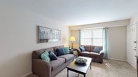 This single-floor residence offers a thoughtfully organized layout with distinct living, dining, and sleeping areas, all finished with wall-to-wall carpeting throughout the main rooms and tile in the kitchen and bathroom. The living room is generously sized, featuring two large sofas, a picture window dressed with teal grommet curtains that allow ample natural light, and tasteful wall art that adds a welcoming ambiance. Adjacent to the living area, a formal dining room provides space for a full-sized dining table and chairs, accented by matching teal curtains and additional wall art. A separate breakfast nook adjacent to the kitchen offers a casual dining option with its own window. The kitchen is equipped with dark wood cabinetry, ample counter space, a double sink, and a full-size refrigerator, with tile flooring for easy maintenance. The bedroom is a restful retreat, featuring carpet flooring, sheer curtains that invite soft natural light, and generous storage provided by a classic wood dresser ensemble. The bathroom is finished with a mosaic-tile floor, a white vanity, and a shower-tub combination. A dedicated entry area with a solid front door completes this comfortable and functional home.