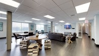 This single-floor space is designed as a multifunctional communal facility, thoughtfully arranged to accommodate both relaxed socializing and practical everyday needs. Throughout the space, drop-panel ceilings with recessed rectangular lighting panels provide consistent, even illumination, complemented by large windows that invite natural light and offer views of the surrounding neighborhood.

The main lounge area is generously proportioned, furnished with dark leather sofas, armchairs, and accent seating including distinctive cowhide-upholstered ottomans and a cowhide area rug. A round wood dining table with cross-leg chairs anchors one end, while teal wingback chairs add a bold accent near the windows. Wall-mounted flat-screen televisions are positioned throughout for easy viewing. Open shelving units and industrial-style décor, including decorative gear wall pieces, contribute to the space's modern yet casual aesthetic.

A dedicated galley-style kitchen runs parallel along one section, featuring light wood cabinetry, granite countertops on both sides, and stainless steel appliances including a refrigerator, gas range, microwave, and dishwasher. Pendant lighting overhead adds a welcoming warmth. Dark tile flooring flows consistently through the lounge areas, unifying the overall design into a cohesive, functional environment.