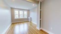This 1-bedroom, 1-bathroom unit offers a clean, modern aesthetic across a single level. The living area is highlighted by warm hardwood flooring that runs throughout, complementing the neutral, light-toned walls that create a bright and welcoming atmosphere. Recessed lighting is installed in the ceiling, providing even, ambient illumination that enhances the sense of openness. A set of double-hung windows allows natural light to pour in, offering views of the surrounding greenery outside. Crisp white baseboards and trim provide a polished contrast to the warm tones of the floors. A distinctive architectural column with built-in decorative shelving adds a unique design element, offering both character and functional display space. The unit also features a compact heating unit tucked neatly into the space, maintaining the clean lines of the room. The overall ambiance is calm and refined, with thoughtful details that balance practicality and style. The neutral palette and quality finishes make this a versatile space well-suited for a variety of living arrangements.
