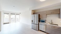This thoughtfully designed single-floor unit offers a clean, modern aesthetic throughout. The open-concept living and kitchen area is bathed in natural light, courtesy of floor-to-ceiling sliding doors that lead to a private balcony, providing pleasant outdoor views and a seamless indoor-outdoor connection. Light wood-tone flooring runs throughout the main living areas, enhancing the bright and airy atmosphere.

The kitchen is a standout feature, outfitted with a full suite of stainless steel appliances including a French-door refrigerator with ice maker, dishwasher, built-in microwave, and a freestanding range. Warm, wood-grain upper and lower cabinetry provides ample storage, complemented by a light countertop and a tiled backsplash that adds texture to the space.

The bathroom is designed with accessibility in mind, featuring sturdy grab bars, a clean white vanity with an integrated sink, tile flooring, and a well-placed mirror. The bedroom offers a cozy and quiet retreat with plush carpeting, neutral-toned walls, and a window that frames views of the surrounding greenery and neighborhood. The overall design palette of whites, creams, and warm wood tones creates a cohesive, welcoming environment throughout the unit.
