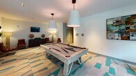 Located at 1743 Central Park Rd in Charleston, South Carolina, this single-floor community amenity space blends modern design with a relaxed coastal aesthetic. The lounge area is anchored by a bold geometric-patterned carpet in shades of teal, beige, and cream, setting a vibrant tone throughout the space. Large floor-to-ceiling windows with dark metal frames flood the seating area with natural light and provide direct views of an outdoor area with lush greenery and a poolside setting, creating a seamless indoor-outdoor connection. Comfortable seating includes a mix of teal and charcoal accent chairs arranged around an eye-catching sculptural wood side table. A separate seating nook features rich leather chairs with gold-toned accents and an artistic living wall display of framed succulent arrangements, adding a natural, textural element to the interior. A full-size billiards table takes center stage in the game room, lit by oversized pendant lights, and accompanied by a wall-mounted television and warm lounge seating for spectators. Adjacent to the lounge, a glass-enclosed fitness center is fully visible through floor-to-ceiling glass panels, housing a variety of cardio and strength equipment. The overall ambiance is contemporary and inviting, designed for both social gatherings and active recreation.