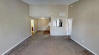 This single-floor residence offers an open and airy layout highlighted by impressive vaulted ceilings that lend a sense of spaciousness to the main living area. The living room is finished with wall-to-wall carpet in a warm, neutral tone, complemented by crisp white baseboards and beige walls that create a clean, inviting atmosphere throughout. A distinctive pass-through window connects the kitchen to the main living space, providing an open sightline and enhancing the flow between the two areas. The kitchen is equipped with standard appliances, including a refrigerator, and features cabinetry that maximizes storage. HVAC vents are positioned near the upper portion of the vaulted walls, ensuring efficient climate control throughout the space. A doorway from the living room leads to additional rooms, offering a functional and well-connected floor plan. The textured ceiling adds a classic residential touch, while the overall neutral palette provides a versatile backdrop that adapts easily to a variety of interior styles. The space is thoughtfully designed to balance openness with defined areas, making it practical for everyday living.