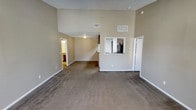 This single-floor residence offers an open and airy layout highlighted by impressive vaulted ceilings that lend a sense of spaciousness to the main living area. The living room is finished with wall-to-wall carpet in a warm, neutral tone, complemented by crisp white baseboards and beige walls that create a clean, inviting atmosphere throughout. A distinctive pass-through window connects the kitchen to the main living space, providing an open sightline and enhancing the flow between the two areas. The kitchen is equipped with standard appliances, including a refrigerator, and features cabinetry that maximizes storage. HVAC vents are positioned near the upper portion of the vaulted walls, ensuring efficient climate control throughout the space. A doorway from the living room leads to additional rooms, offering a functional and well-connected floor plan. The textured ceiling adds a classic residential touch, while the overall neutral palette provides a versatile backdrop that adapts easily to a variety of interior styles. The space is thoughtfully designed to balance openness with defined areas, making it practical for everyday living.