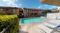This residential complex showcases a classic Southern California architectural style, characterized by warm tan stucco exteriors, distinctive red clay tile roofing, and clean geometric lines across its two-story structure. The buildings are arranged to frame a central outdoor courtyard, where a generously sized swimming pool serves as the focal point of the community amenity space. The pool area is surrounded by a concrete deck furnished with lounge chairs and a shaded patio area supported by textured stucco columns, providing a comfortable space for relaxation under open skies. Lush greenery, including hedges and tropical plantings such as banana trees, softens the hardscape and adds a vibrant touch to the outdoor environment. Wrought iron fencing encloses the pool area, offering both security and an open, airy feel. The upper-level units are accessible via open-air walkways lined with metal railings, which receive ample natural light through the day as sunlight casts distinctive shadow patterns across the corridor floors. Individual unit entry doors are finished in a muted tone that complements the neutral stucco walls. The overall ambiance of the complex is relaxed and sun-drenched, reflecting a laid-back residential lifestyle well-suited to a warm climate.