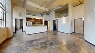 Located at 675 North Main Street in downtown Winston-Salem, this urban loft showcases a distinctive industrial character. The open-plan living and kitchen area features polished concrete floors, soaring ceilings with exposed ductwork, and structural columns that highlight the building's heritage. The kitchen includes dark wood cabinetry, stainless steel appliances, and a breakfast bar. Large factory-style windows flood the space with natural light and frame city skyline views. The generous bedroom offers tall windows with a ceiling fan. Both bathrooms are finished with tile flooring and tub-shower combinations. A dedicated laundry room and hallway complete the layout.