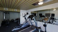 This fitness center is a purpose-built exercise space designed to provide a comprehensive workout environment within a compact footprint. The room is outfitted with a selection of cardio equipment, including a treadmill and an elliptical trainer, allowing users to engage in a variety of aerobic exercises. A free weight area with medicine balls and a weight rack rounds out the strength training options. Full-length mirrors line one wall, creating a sense of depth and allowing users to monitor their form during workouts. The ceiling features a distinctive striped pattern in muted gold and green tones, adding a unique visual character to the space. Overhead flush-mount lighting ensures the room is well-lit throughout. Ventilation is supported by ceiling vents, maintaining a comfortable environment during exercise. The white walls keep the space feeling clean and open, while the dark flooring provides a practical, durable surface suited for gym activity. Overall, the fitness center is thoughtfully arranged to maximize utility and accessibility, making it a functional and inviting space for daily exercise.