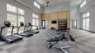 Located at 174 Overlook Point Place in Charleston, SC, this well-appointed fitness center offers a comprehensive workout environment designed to accommodate a wide range of exercise routines. The single-floor facility boasts soaring ceilings that lend an open, airy feel, while an abundance of large windows along the perimeter floods the space with natural light and provides views of the surrounding community.

The main workout area features a row of TRUE-brand treadmills and cardio machines positioned along the windowed walls, complemented by a set of stationary cycling bikes for group or individual spin sessions. A dedicated strength training zone includes a multi-station cable machine, a full dumbbell rack ranging across multiple weight increments, kettlebells, and medicine balls organized on a bold blue storage rack. Additional functional training equipment, including benches and specialty attachments, rounds out the strength offerings.

Full-length mirrors line one wall, enhancing the sense of space and supporting proper form during workouts. The flooring throughout features a distinctive checkered carpet tile pattern in neutral grey tones, providing both comfort and durability. Ceiling fans maintain airflow across the space, while a wall-mounted flat-screen television adds an entertainment option. A textured accent wall adds a touch of design character, creating an inviting atmosphere that balances functionality with modern aesthetics.