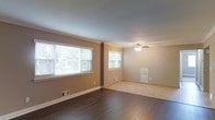 This single-story home offers a welcoming and functional layout designed for comfortable living. The main living area is a standout feature, showcasing rich, dark hardwood flooring that adds warmth and elegance to the space. Multiple large windows allow an abundance of natural light to fill the room, creating an open and airy atmosphere throughout the day. The neutral, taupe-toned walls complement the flooring and provide a versatile backdrop for a variety of interior styles.

A transition from the hardwood floor to light-toned carpeting delineates the adjoining room, which is fitted with a classic ceiling fan with integrated lighting — a practical and timeless fixture. Crown molding details along the ceiling add a refined architectural touch that elevates the overall aesthetic of the interior.

The space benefits from consistent, neutral tones across both rooms, contributing to a cohesive and calming ambiance. Floor vents are positioned throughout, indicating a central HVAC system for year-round climate control. The generous window placement not only enhances natural illumination but also offers views of the surrounding greenery, connecting the interior to the outdoors. Overall, this home presents a clean, well-maintained, and inviting environment.