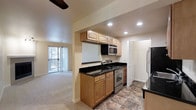 This single-story space is designed with a functional, open layout that connects the kitchen and living areas seamlessly. The kitchen is outfitted with warm honey-toned wood cabinetry, both upper and lower, providing ample storage. Dark granite countertops add a sleek contrast to the light cabinetry, while stainless steel appliances — including a range, built-in microwave, and refrigerator — lend a modern touch. Recessed lighting illuminates the kitchen workspace effectively. The flooring in the kitchen features a durable stone-look tile, transitioning into plush carpeting in the adjacent living area. The living space is brightened by a sliding glass door that opens to a private balcony, allowing natural light to filter in and offering outdoor access. Vertical blinds provide light control and privacy. A ceiling fan is positioned overhead in the living area, adding to the comfort of the space. A hallway visible from the kitchen suggests additional rooms beyond, and the overall layout is practical and well-organized, making efficient use of the available square footage.