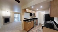 This single-story space is designed with a functional, open layout that connects the kitchen and living areas seamlessly. The kitchen is outfitted with warm honey-toned wood cabinetry, both upper and lower, providing ample storage. Dark granite countertops add a sleek contrast to the light cabinetry, while stainless steel appliances — including a range, built-in microwave, and refrigerator — lend a modern touch. Recessed lighting illuminates the kitchen workspace effectively. The flooring in the kitchen features a durable stone-look tile, transitioning into plush carpeting in the adjacent living area. The living space is brightened by a sliding glass door that opens to a private balcony, allowing natural light to filter in and offering outdoor access. Vertical blinds provide light control and privacy. A ceiling fan is positioned overhead in the living area, adding to the comfort of the space. A hallway visible from the kitchen suggests additional rooms beyond, and the overall layout is practical and well-organized, making efficient use of the available square footage.