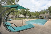 This outdoor pool area offers a well-designed recreational space ideal for relaxation and leisure. The centerpiece is a generously sized rectangular swimming pool with clear, blue water, accessible via a metal entry railing. The surrounding concrete deck provides ample space for lounging, furnished with a collection of reclining chairs and canopied swing loungers in teal, offering both sun and shade options for visitors.

A dedicated seating area features a metal mesh table and chairs, providing a comfortable spot for outdoor dining or socializing. The entire pool area is enclosed by a sturdy black iron fence with pointed finials, ensuring both security and a clean, classic aesthetic. A gated entry controls access to the space.

The backdrop is defined by a dense canopy of mature trees, including towering pines and broad-leafed varieties draped with natural moss, creating a lush, shaded perimeter that enhances privacy and provides a natural aesthetic. Residential buildings are visible beyond the fencing, situating this amenity within a broader community setting. The space benefits from abundant natural light under open skies, making it an inviting outdoor retreat during sunny days.