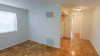 This single-floor space showcases warm parquet wood flooring that extends seamlessly throughout the main living area, hallway, and into the adjacent spaces, adding a timeless character to the interior. The living area is finished with clean, white walls that provide a bright, neutral backdrop, complemented by flush-mounted ventilation grilles for climate control. A partial dividing wall separates the living space from the kitchen area, which is visible beyond and features light wood cabinetry and a white refrigerator. The hallway, illuminated by a ceiling-mounted light fixture, connects the main areas of the unit and leads toward what appears to be additional rooms. The overall layout is straightforward and functional, with a consistent flooring material tying the spaces together cohesively. The white walls and light flooring contribute to a clean, well-lit ambiance throughout. The space offers a practical, no-frills design that provides a solid foundation for personalization and furnishing.