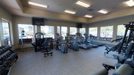 This fitness center offers a bright and functional workout environment designed to support a range of exercise routines. Large windows line multiple walls, flooding the space with natural light and providing open views of the surrounding exterior. The room is finished with neutral-toned carpet tiles and a smooth ceiling fitted with recessed fluorescent lighting, ensuring consistent illumination throughout.

The cardio section is prominently featured along the windowed wall and includes a row of treadmills and an elliptical trainer, all positioned to take advantage of the outdoor views. A cable machine and strength training station anchor one side of the room, catering to resistance and weight-based workouts. Dumbbells are also available, adding versatility for free-weight exercises. Two wall-mounted flat-screen televisions are positioned at either end of the space, providing entertainment during workouts. The layout is organized and spacious, allowing for comfortable movement between equipment. Overall, the fitness center presents a clean, well-maintained environment suitable for both cardio and strength training.
