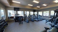 This fitness center offers a bright and functional workout environment designed to support a range of exercise routines. Large windows line multiple walls, flooding the space with natural light and providing open views of the surrounding exterior. The room is finished with neutral-toned carpet tiles and a smooth ceiling fitted with recessed fluorescent lighting, ensuring consistent illumination throughout.

The cardio section is prominently featured along the windowed wall and includes a row of treadmills and an elliptical trainer, all positioned to take advantage of the outdoor views. A cable machine and strength training station anchor one side of the room, catering to resistance and weight-based workouts. Dumbbells are also available, adding versatility for free-weight exercises. Two wall-mounted flat-screen televisions are positioned at either end of the space, providing entertainment during workouts. The layout is organized and spacious, allowing for comfortable movement between equipment. Overall, the fitness center presents a clean, well-maintained environment suitable for both cardio and strength training.