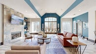 Located at 3209 Westbury Lake Dr in Charlotte, NC, this thoughtfully designed single-floor living space offers a welcoming and modern atmosphere defined by bold design choices and versatile functionality. The open-concept layout centers around a spacious living room that immediately draws attention with its dramatic vaulted ceilings, which are accentuated by striking teal architectural beams and accent walls that add vivid personality to the space.

Natural light pours in through large arched and floor-to-ceiling windows, illuminating light wood-tone flooring that runs throughout. A floor-to-ceiling stone tile fireplace serves as a stunning focal point, flanked by open floating shelves that offer both display and functional storage. Adjacent to the fireplace, a dedicated nook with a built-in desk and shelving provides a compact workspace.

The living area is further enhanced by a stylish seating arrangement featuring a rich cognac leather sofa, mid-century modern accent chairs, a round wood coffee table, and a geometric patterned area rug. On the opposite end, a kitchen bar with pendant lighting and industrial-style barstools connects seamlessly to a kitchen equipped with dark wood cabinetry and stainless steel appliances. The overall ambiance is vibrant, modern, and inviting — ideal for both relaxing and social gatherings.