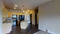 This single-floor space showcases an open-concept layout that seamlessly connects the kitchen and living areas. The kitchen is equipped with light wood cabinetry, black appliances including a refrigerator and range, and a central island topped with a dark granite-style countertop that doubles as a breakfast bar. Pendant lights hang above the island, complementing the recessed lighting throughout the ceiling to create a well-lit environment. Dark hardwood flooring runs through the main living and kitchen areas, providing a warm contrast to the neutral-toned walls. Transitioning toward the living space, the flooring shifts to carpet, visually defining the separate zones within the open floor plan. Beyond the kitchen, a hallway leads to what appears to be a spacious walk-in closet with built-in shelving, alongside a separate storage or utility closet. The layout is practical and efficient, with clear sightlines from the kitchen through to adjacent rooms. The overall ambiance is modern and functional, with clean architectural lines and thoughtful storage solutions integrated throughout the space.
