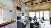 This single-floor space presents an elegantly designed lounge area that combines architectural sophistication with comfortable, contemporary furnishings. The room's most commanding feature is a floor-to-ceiling stacked stone fireplace, complete with a classic white mantel and a wall-mounted flat-screen television above, serving as a natural focal point for the space. The soaring ceilings are enhanced by rich, dark wood coffered detailing, adding a layer of refined architectural interest overhead. Soft blue-gray walls create a calm, neutral backdrop that complements the warm tones of the stone and woodwork throughout. Natural light flows generously into the space through large windows dressed with patterned Roman shades, offering views of the surrounding greenery outside. The seating arrangement includes sleek dark leather sofas and chairs accented with decorative pillows in yellow, cream, and patterned fabrics, centered around a modern glass and chrome coffee table. Additional seating near the windows provides intimate conversation areas. Decorative artwork and tall sculptural vases complete the polished, welcoming aesthetic of this well-appointed gathering space.