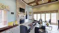 This single-floor space presents an elegantly designed lounge area that combines architectural sophistication with comfortable, contemporary furnishings. The room's most commanding feature is a floor-to-ceiling stacked stone fireplace, complete with a classic white mantel and a wall-mounted flat-screen television above, serving as a natural focal point for the space. The soaring ceilings are enhanced by rich, dark wood coffered detailing, adding a layer of refined architectural interest overhead. Soft blue-gray walls create a calm, neutral backdrop that complements the warm tones of the stone and woodwork throughout. Natural light flows generously into the space through large windows dressed with patterned Roman shades, offering views of the surrounding greenery outside. The seating arrangement includes sleek dark leather sofas and chairs accented with decorative pillows in yellow, cream, and patterned fabrics, centered around a modern glass and chrome coffee table. Additional seating near the windows provides intimate conversation areas. Decorative artwork and tall sculptural vases complete the polished, welcoming aesthetic of this well-appointed gathering space.