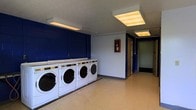 Located at 4645 Tamarack Blvd in Columbus, Ohio, this on-site laundry facility offers a practical and functional space designed to serve building residents efficiently. The single-floor room is equipped with a row of commercial-grade front-loading washing machines and dryers lined along a bold navy blue accent wall, providing ample capacity for laundry needs. The flooring is a durable, easy-to-clean light vinyl, well-suited for a high-traffic utility space. Overhead fluorescent lighting illuminates the room evenly, ensuring a bright and comfortable environment. A fire extinguisher is prominently mounted on the wall, reflecting attention to safety standards. A doorway at the rear of the room provides additional access, contributing to a practical flow within the space. The overall layout is straightforward and spacious enough to allow multiple users to move comfortably. The combination of functional equipment, safety features, and clean design makes this laundry facility a convenient and reliable amenity for residents of the property.