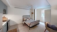 This efficiently designed single-floor unit makes smart use of its compact footprint, offering a thoughtfully arranged open-concept layout that integrates the living, sleeping, and kitchen areas into one cohesive space. Light wood-toned flooring runs throughout, lending warmth and visual continuity across the unit.

The spacious primary bedroom occupies the majority of the floor plan and features a ceiling fan, a window that brings in natural light, and ample room for a full bed and workspace. A dedicated work area with a desk and seating provides functional versatility within the bedroom zone.

The kitchen is positioned along one wall and is outfitted with slate-gray cabinetry, stainless steel appliances including a refrigerator, stove, and microwave, as well as a sink with under-counter storage. A ceiling fan overhead aids in air circulation across the open living and kitchen area.

In-unit stacked washer and dryer are tucked neatly into a recessed alcove, adding a practical convenience. The bathroom features light-toned walls, wood-style flooring, a shower with curtain, and matte black fixtures. A hallway with wall-mounted storage hooks and shelving connects the entry to the main living space, maximizing organization in a compact setting.
