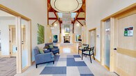 This well-appointed clubhouse in Loveland, Colorado offers a generous single-floor layout thoughtfully designed for community gathering and recreation. The centerpiece is a grand main lounge showcasing soaring vaulted ceilings with exposed wood beams, warm hardwood flooring, and an impressive stone fireplace. Floor-to-ceiling windows and clerestory glazing flood the space with natural light, while French doors open directly to an outdoor patio area. A dedicated game room features a full-size pool table, foosball, an arcade cabinet, dartboard, and comfortable seating beneath high ceilings, creating an energetic social hub. The private theater room is equipped with tiered seating — including plush recliners and lounge sofas — a ceiling-mounted projector, and a large projection screen framed by acoustic wall panels for an immersive viewing experience. A café-style lounge provides a beverage station, dining tables, and wall-mounted TV, ideal for casual gatherings. The conference room offers a long boardroom table surrounded by upholstered chairs beneath a statement pendant light, accented by colorful artwork and generous natural light. Throughout, patterned carpeting, warm wood trim, and a cohesive modern palette tie the spaces together into a versatile and inviting community destination.