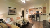 This single-floor residence offers a thoughtfully designed open-concept layout that seamlessly connects the living, dining, and kitchen areas into one cohesive and functional space. The living area is warm and inviting, featuring rich hardwood flooring, crown molding, and a ceiling fan with integrated lighting that adds both comfort and style. Neutral-toned walls are complemented by warm accent lighting, creating an intimate yet airy ambiance throughout.

The transition into the dining area is effortless, where a bistro-style high-top table and bar seating provide a casual yet elegant setting for meals and entertaining. A bold terracotta accent wall serves as a striking focal point, adding a pop of color and visual depth to the open layout.

The kitchen is well-equipped with light wood cabinetry, dark countertops, and black appliances, including a built-in microwave. Track lighting overhead ensures the space is well-lit and functional. A decorative pendant light adds a refined touch above the kitchen area.

Throughout the space, carefully chosen décor — including framed wall art, floral centerpieces, and textured soft furnishings — contributes to an overall aesthetic that is both stylish and livable. The open sightlines across all areas enhance the sense of spaciousness, making this a comfortable and visually appealing home.