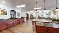 This thoughtfully designed single-floor community space seamlessly blends functional elegance with a warm, welcoming atmosphere. The layout is anchored by a generously sized kitchen outfitted with rich cherry wood cabinetry, dark granite countertops, and stainless steel appliances, including a double wall oven and dishwasher. Vibrant, handcrafted decorative tile work serves as a striking backsplash, adding an artistic focal point to the kitchen area. A central island with turned wood legs and a stainless steel drawer unit provides additional workspace and a natural gathering point. Hardwood flooring flows throughout, lending warmth and continuity across the space. Elegant arched doorways and ornate wrought-iron decorative elements contribute a distinctive architectural character. The adjacent dining area features soaring ceilings with coffered detailing, tall windows dressed with blinds that allow generous natural light while offering privacy, and a bold patterned carpet that grounds the dining furniture. Tables and chairs are arranged to accommodate multiple dining groups comfortably. Framed artwork adorns the walls, and lush potted palms add natural greenery. Stately columns and sets of glass-paned French doors elegantly delineate the transition between the kitchen and dining areas, creating a cohesive and inviting environment throughout.