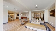 The Los Alisos at Mission Viejo Clubhouse is a thoughtfully designed 1,280 square foot community facility located in Mission Viejo, CA. The single-floor layout encompasses distinct functional zones that flow seamlessly into one another, all unified by light wood-tone flooring and a warm, neutral color palette of whites and soft beiges.

The main lounge area serves as the centerpiece of the clubhouse, featuring a large sectional sofa, woven accent chairs, and a wall-mounted flat-screen TV set within a recessed niche. Bold black-and-white geometric rugs anchor the seating arrangements, while recessed lighting provides a clean, modern ambiance. Sliding glass doors allow natural light to fill the space and offer views to the exterior.

Adjacent to the lounge is a dining area with a marble-topped table, modern pendant lighting, and a large framed mirror that visually expands the room. A dedicated conference/co-working room accommodates several guests around a light wood table, complemented by wall-mounted display screens and framed abstract artwork.

The kitchenette features stone countertops, warm wood cabinetry with brushed gold hardware, a stainless steel sink with a gold gooseneck faucet, and a stone backsplash — combining functionality with refined, upscale finishes throughout.