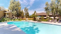 Located at Arroyo Park Drive in Santa Clarita, California, this outdoor amenity space offers a well-appointed resort-style recreation area designed for leisure and relaxation. The centerpiece is a generously sized swimming pool with a gently curved shape, featuring convenient entry steps and a shallow wading area, all maintained with clear, inviting water. Adjacent to the main pool is a circular spa, nestled within a naturalistic rock garden setting complete with boulders and lush ornamental plantings that create a tranquil, park-like atmosphere. The surrounding pool deck provides ample space for sunbathing, furnished with multiple lounge chairs and shaded seating areas with patio umbrellas. Towering mature pine and shade trees frame the entire area, offering natural shade and a sense of seclusion. A covered pavilion structure with a pitched roofline serves as a poolside facility building, providing convenient access to amenities. Landscaped pathways wind through native-style plantings, stone accents, and greenery, connecting various areas of the outdoor space. The overall ambiance is bright and sun-filled, reflecting the characteristic Southern California climate, and the space is enclosed by perimeter fencing for privacy and security.