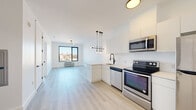 This contemporary single-floor unit showcases a clean, modern design throughout, highlighted by light hardwood-style flooring and crisp white walls that create a bright, welcoming atmosphere. The open-concept layout seamlessly connects the kitchen and living area, maximizing the sense of space and encouraging a natural flow between functional zones.

The kitchen is well-appointed with a full suite of stainless steel appliances, including an electric range, over-the-range microwave, dishwasher, and refrigerator. Sleek white cabinetry extends to the upper walls, offering generous storage, while light-toned countertops and a matte black faucet at the sink add a polished contrast. A stylish multi-bulb pendant light fixture hangs above, enhancing the modern aesthetic.

The bedroom features large, black-framed windows that frame elevated city views, flooding the room with natural light. A wall-mounted PTAC heating and cooling unit ensures year-round comfort. The open living area also benefits from city-facing windows and a contemporary light fixture overhead.

The bathroom is finished with white subway tile in the walk-in shower, complemented by matte black fixtures and hardware, maintaining a cohesive modern style. Throughout the unit, the consistent use of neutral tones, contemporary fixtures, and thoughtful design details come together to deliver a polished and functional living space.