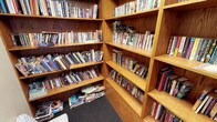 This inviting single-floor space serves as a well-stocked library or reading room, designed with both functionality and warmth in mind. The room is defined by its built-in, floor-to-ceiling wooden bookshelves crafted from rich, warm-toned oak or similar hardwood, lending the space a classic, scholarly ambiance. The shelving units are arranged in a corner configuration, maximizing storage capacity and creating an enveloping, immersive environment for book lovers.

The collection housed within is notably diverse, spanning a wide range of genres including fiction, non-fiction, cookbooks, and more, with titles organized across multiple shelves. The sheer volume of books — many tightly packed and some stacked horizontally — speaks to the breadth and depth of the collection available. The shelves are filled to capacity, reflecting a well-used and cherished reading resource.

The flooring features a dark, textured carpet that adds a quiet, comfortable feel underfoot and complements the natural tones of the woodwork. The overall atmosphere of the space is warm, intimate, and intellectually stimulating — ideal for browsing, reading, or quiet study. The corner shelving design creates a sense of being surrounded by knowledge, making this a standout feature of the space.
