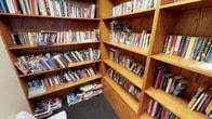 This inviting single-floor space serves as a well-stocked library or reading room, designed with both functionality and warmth in mind. The room is defined by its built-in, floor-to-ceiling wooden bookshelves crafted from rich, warm-toned oak or similar hardwood, lending the space a classic, scholarly ambiance. The shelving units are arranged in a corner configuration, maximizing storage capacity and creating an enveloping, immersive environment for book lovers.

The collection housed within is notably diverse, spanning a wide range of genres including fiction, non-fiction, cookbooks, and more, with titles organized across multiple shelves. The sheer volume of books — many tightly packed and some stacked horizontally — speaks to the breadth and depth of the collection available. The shelves are filled to capacity, reflecting a well-used and cherished reading resource.

The flooring features a dark, textured carpet that adds a quiet, comfortable feel underfoot and complements the natural tones of the woodwork. The overall atmosphere of the space is warm, intimate, and intellectually stimulating — ideal for browsing, reading, or quiet study. The corner shelving design creates a sense of being surrounded by knowledge, making this a standout feature of the space.