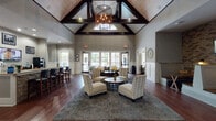 This single-floor amenity and clubhouse space combines sophisticated design with a welcoming, community-focused atmosphere. The centerpiece is a grand main lounge featuring soaring vaulted ceilings with striking dark wood beam accents and shiplap detailing, anchored by a statement chandelier that draws the eye upward. Rich hardwood flooring runs throughout, adding warmth and continuity across the space.

The main lounge area is thoughtfully furnished with accent chairs, plush seating, and a large area rug, creating an inviting gathering space. Large floor-to-ceiling windows and glass doors line the rear wall, flooding the interior with natural light and offering direct views of the outdoor pool and surrounding greenery. A cozy banquette nook, accented by exposed brick and wood-paneled walls, provides a more intimate setting complete with a wall-mounted television.

A well-appointed kitchenette/bar area features granite countertops, dark cabinetry, a sink, under-counter refrigeration, and a coffee station, making it ideal for social gatherings. Adjacent bar-height seating at granite-topped islands adds to the communal feel. A dedicated study room offers built-in granite desks and office chairs for focused work. The entry corridor features wainscoting and decorative details that set an elegant tone from arrival.