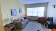 Located at 951 Route 13 in Cortland, New York, this single-floor space offers a comfortable and functional layout designed for everyday living. The living area is the heart of the space, featuring soft, warm yellow-toned walls that create a bright and welcoming atmosphere. A large multi-pane window spans a generous portion of one wall, allowing abundant natural light to fill the room and offering views of the surrounding greenery outside. The living area is thoughtfully arranged with a plush sofa accented by decorative pillows and complemented by a cozy armchair, both in warm earthy tones. A soft area rug anchors the seating arrangement, adding texture and warmth to the space. Tasteful wall art in cool blue tones provides a pleasing contrast to the warm wall color, contributing to a balanced and calming aesthetic. A small side table with a lamp adds a subtle, warm glow to the room. The overall ambiance is inviting and relaxed, with a clean, uncluttered design that emphasizes comfort and livability. The space is well-suited for those seeking a peaceful, move-in-ready environment in the Cortland, NY area.
