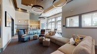 This thoughtfully designed community clubhouse delivers a sophisticated blend of comfort and functionality across a single, open floor. Soaring ceilings define the main lounge, where dramatic wood-paneled tray ceilings and oversized ring pendant lights create a striking focal point overhead. Large floor-to-ceiling windows flood the space with natural light and offer views of the surrounding outdoor pool area and grounds, enhancing the bright, welcoming atmosphere.

The main lounge is generously furnished with plush sectional sofas, accent chairs, and low coffee tables arranged to encourage both relaxed socializing and quiet retreat. Adjacent to the lounge, a sleek wet bar features a light-colored countertop with bar stool seating, backed by warm yellow subway tile and cascading Edison-style pendant lights, adding an inviting, upscale touch. A foosball table nearby adds a playful recreational element to the space.

A separate seating area with exposed wood ceiling beams and a starburst chandelier provides a cozier, intimate ambiance, while a glass-enclosed conference room with a round table and modern chairs offers a dedicated space for meetings or focused work. A built-in co-working station with a live-edge wood table and multiple seating positions rounds out the functional amenities, all framed by views of the exterior through a row of windows.