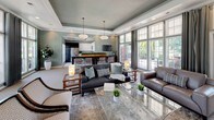 Located at 1011 W Butler Rd in Mauldin, South Carolina, this single-floor community clubhouse offers a thoughtfully designed gathering space with a refined, contemporary aesthetic. The interior is defined by soft sage-green walls, elegant tray ceilings with crown molding, and recessed lighting complemented by modern pendant fixtures that create a warm, inviting ambiance throughout.

The main lounge area is generously proportioned, furnished with a mix of leather and upholstered sofas, accent chairs, and glass-topped coffee tables arranged to encourage both relaxed socializing and casual conversation. Large floor-to-ceiling windows and multiple sets of French doors flood the space with natural light while providing direct views of the outdoor pool area.

A long, dark-topped bar-height counter with cushioned bar seating serves as a central feature, functioning as a communal gathering and dining area. Adjacent to this is a well-appointed kitchenette with stainless steel appliances and white cabinetry. A dedicated workspace nook is tucked into one corner, complete with a built-in desk and wall-mounted television. A classic white fireplace with decorative surround anchors another corner of the space, adding a cozy focal point. Bold artwork and lush greenery accents complete the polished, welcoming atmosphere.