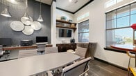 This well-appointed conference and business center room offers a sophisticated and functional environment designed for productivity and collaboration. The space is anchored by a large central conference table surrounded by cushioned rolling chairs, accommodating multiple attendees comfortably. High ceilings with bold dark crown molding add architectural interest and a sense of openness, while multiple windows flood the room with natural light, offering views to the outdoors.

One wall features a dedicated workstation counter lined with desktop computers and a printer, making it ideal for business center use. A textured dark accent wall serves as a striking backdrop for this workstation area, complemented by decorative metallic disc wall art and an industrial-style pendant light. Opposite, built-in dark wood cabinetry with a wall-mounted flat-screen TV and open shelving displaying decorative accents provide both storage and visual appeal.

Elegant French doors with dark frames open to an adjacent common area, enhancing connectivity and visibility throughout the facility. A large mirror near an arched doorway reflects light and adds depth. A cozy nook with a loveseat, golden ottomans, and a wall-mounted TV offers a relaxed alternative seating option. Patterned carpet flooring ties the design together, creating a cohesive, modern aesthetic throughout.