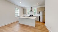 Located in Nashville, TN, this well-appointed three-story townhome offers a clean, modern aesthetic throughout. The main living level features an open-concept layout where the living area flows seamlessly into a bright, contemporary kitchen. The kitchen is outfitted with grey shaker-style cabinetry, white quartz countertops, a subway tile backsplash, stainless steel appliances including a range with a stainless hood vent and refrigerator, and a center island with a built-in sink — all illuminated by natural light from multiple windows overlooking lush greenery. A convenient half bath with a pedestal sink and oval mirror is also found on this level, finished with light hardwood-style flooring that runs throughout the main areas. The upper floor houses the bedrooms, each carpeted for comfort and featuring closets with sliding or hinged panel doors. A full bathroom with a tub-shower combo, white vanity, and light hardwood-style flooring serves the upper level. A stacked Whirlpool washer and dryer unit is tucked into a dedicated laundry closet near the stairwell for easy access. The ground floor includes an attached garage with a coffered ceiling, recessed lighting, and an automatic opener, providing ample parking and storage space.