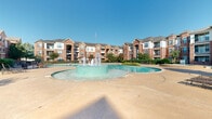 Located at 9000 Almeda Rd in Houston, Texas, this outdoor amenity area is the centerpiece of a multi-story brick residential community. The space is anchored by a generously sized, freeform swimming pool with vivid blue water, complemented by a dedicated circular fountain feature with a tiered water jet that creates a striking visual focal point. Adjacent to the main pool are two raised spa/hot tub basins, each accessed by curved concrete steps, offering a more intimate retreat within the broader outdoor setting.

The pool deck is finished in warm-toned stamped concrete, providing ample space for lounging and circulation. Cushioned lounge chairs and seating clusters are thoughtfully distributed throughout, including a shaded pergola area outfitted with outdoor dining furniture for al fresco gatherings. Mature trees and manicured hedgerows frame the perimeter, delivering shade and a sense of privacy while lush ornamental plantings add color and texture throughout the landscape.

The surrounding multi-story brick buildings feature private balconies that overlook the amenity courtyard, creating an enclosed, resort-like atmosphere. Under the expansive Texas sky, this outdoor space is designed to provide residents with a comprehensive and well-appointed recreational environment year-round.