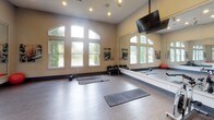 This single-level fitness facility is thoughtfully designed to accommodate a wide range of exercise activities. The space is divided into distinct functional zones, beginning with a spacious group exercise studio that immediately impresses with its soaring ceilings and dramatic arched windows that flood the room with natural light and offer views of surrounding greenery. Full-length wall mirrors amplify the sense of openness and allow participants to monitor their form during workouts. The studio is equipped with exercise mats, medicine balls, a stability ball, and a dumbbell rack, making it well-suited for yoga, stretching, and group fitness classes. A wall-mounted flat-screen television provides additional functionality for guided workout sessions. The flooring transitions between hardwood-style planks in the studio zone and carpet in adjacent areas, creating visual distinction between spaces. A secondary area accommodates cardio equipment, including stationary bikes and treadmills, visible through sleek glass-paneled double doors that connect the zones seamlessly. Recessed lighting throughout ensures consistent illumination, while motivational wall art adds personality to the neutral, warm-toned walls. The overall ambiance is clean, modern, and energizing, providing an inviting environment for a comprehensive fitness experience.