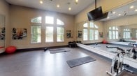 This single-level fitness facility is thoughtfully designed to accommodate a wide range of exercise activities. The space is divided into distinct functional zones, beginning with a spacious group exercise studio that immediately impresses with its soaring ceilings and dramatic arched windows that flood the room with natural light and offer views of surrounding greenery. Full-length wall mirrors amplify the sense of openness and allow participants to monitor their form during workouts. The studio is equipped with exercise mats, medicine balls, a stability ball, and a dumbbell rack, making it well-suited for yoga, stretching, and group fitness classes. A wall-mounted flat-screen television provides additional functionality for guided workout sessions. The flooring transitions between hardwood-style planks in the studio zone and carpet in adjacent areas, creating visual distinction between spaces. A secondary area accommodates cardio equipment, including stationary bikes and treadmills, visible through sleek glass-paneled double doors that connect the zones seamlessly. Recessed lighting throughout ensures consistent illumination, while motivational wall art adds personality to the neutral, warm-toned walls. The overall ambiance is clean, modern, and energizing, providing an inviting environment for a comprehensive fitness experience.