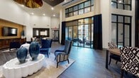 This sophisticated single-story space showcases a bold contemporary design anchored by soaring double-height ceilings that amplify the sense of openness throughout. The main lounge area centers on a sculptural white scalloped coffee table surrounded by deep navy blue wingback chairs and patterned accent seating, all set atop a neutral area rug over wide-plank wood-look tile flooring. A dramatic oversized bronze pendant light commands attention overhead, complementing a coffered ceiling detail finished in dark charcoal.

Floor-to-ceiling glass walls with black steel framing line one side of the space, allowing generous natural light to pour in and offering direct views of an outdoor patio area. Long draping curtains in navy and cream frame the expansive glazing. A dedicated lounge corner features a warm wood-paneled media wall housing a flat-screen television, flanked by upholstered seating and a floor lamp.

Adjacent to the main seating area, a kitchen and dining zone is outfitted with sleek cabinetry, stainless steel appliances, a white tile backsplash, and stylish globe pendant lighting. Curated décor throughout—including lunar phase photography, a starburst mirror, a geometric-patterned credenza, and a gold sunburst mirror—adds layers of visual interest and a cohesive, design-forward aesthetic.
