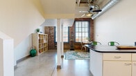 Located in Cleveland's vibrant Superior Ave corridor, this two-bedroom, two-floor loft unit masterfully combines industrial heritage with contemporary comfort. The main level features polished concrete floors, exposed brick walls, and prominent ductwork that lend an authentic urban character. Large, grid-paned windows flood the open living and dining area with natural light, while a ceiling fan with exposed spiral ductwork adds to the industrial aesthetic. A peninsula kitchen anchors the space with crisp white cabinetry, dark stone countertops, a mosaic tile backsplash, and stainless steel appliances — functional and stylish in equal measure. A long hardwood-floored corridor connects the main living area to the carpeted staircase leading to the upper level. Upstairs, the expansive primary bedroom offers soaring ceilings with exposed structural beams, plush carpeting, and a well-sized closet with built-in shelving. The full bathroom features warm terracotta tile floors, a granite-topped vanity with white cabinetry, mosaic tile accents, and a tub-shower combination. A second bedroom provides additional flexible living space. Throughout, the layering of raw industrial elements with clean, modern finishes creates a distinctive and inviting urban retreat.