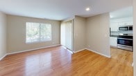 This single-floor residence offers a thoughtfully designed layout with consistent warm-toned hardwood-style flooring running throughout every room, creating a cohesive and inviting atmosphere. The kitchen is a standout feature, showcasing crisp white shaker cabinetry, granite countertops, and a full suite of stainless steel appliances including a refrigerator, electric range, over-the-range microwave, and dishwasher. A undermount sink and ample counter space make this kitchen as functional as it is attractive. The living area is generously proportioned, bathed in natural light from large windows dressed with vertical blinds that offer views of lush greenery outside. A second bright room with a large window provides flexibility as a bedroom or additional living space. The primary bedroom features direct access to a walk-in closet with built-in shelving, as well as a private bathroom connection. A secondary bedroom includes a reach-in closet with a bifold door. The bathroom is finished with clean white tile surrounding a full soaking tub and shower combination. A hallway connects all areas of the home efficiently, and the overall neutral color palette of warm beige walls and white trim lends the space a clean, move-in ready appeal.
