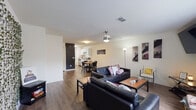 This single-floor unit offers a practical and comfortable open-concept layout, with hardwood-style flooring running throughout the main living areas to create a cohesive, unified feel. The spacious living room is a central gathering space, furnished with two black leather sofas centered around a wooden coffee table. A large flat-screen TV is mounted on the wall, and a wide window with vertical blinds allows natural light to fill the room. A decorative accent wall adorned with trailing faux greenery adds a distinctive visual element, complementing the unit's relaxed aesthetic. The living area flows naturally into the dining space, where a round table and chairs provide a cozy spot for meals. A wall-mounted mirror near the entryway and a chalkboard-painted accent wall near the front door add character to the transitional spaces. The galley-style kitchen is clean and functional, featuring white cabinetry, light stone-look countertops, stainless steel appliances including a range, microwave, and dishwasher, and a stainless steel sink. A rear door in the kitchen provides additional access. Ceiling fans with integrated lighting fixtures are present in the main living areas, ensuring comfortable airflow throughout the unit.