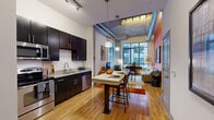This 1-bedroom, 1-bathroom residence in Baltimore's 21231 zip code showcases a compelling loft-inspired aesthetic that blends industrial character with modern finishes. Warm bamboo hardwood flooring runs throughout the open-plan living and kitchen areas, anchoring the space with a cohesive, contemporary feel. The kitchen is well-appointed with dark espresso cabinetry, granite countertops, a full suite of stainless steel appliances, tile backsplash, and a large granite-topped island that doubles as a dining area with bar seating. The open living area is distinguished by soaring ceilings with exposed ductwork and concrete elements, track lighting, and expansive floor-to-ceiling windows that flood the space with natural light and urban views. A bold accent wall in deep orange adds a striking focal point to the living room. The bedroom features a vivid cobalt blue accent wall, plush carpeting, large windows, and exposed concrete ceiling details that maintain the industrial theme. A spacious walk-in closet with full wire shelving and hanging rods provides generous storage. The bathroom is finished with large-format tile flooring, dark vanity cabinetry, white countertops, and a tiled tub/shower combination.