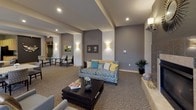 This thoughtfully designed single-floor community space offers a cohesive blend of functionality and refined comfort. The open floor plan encompasses several distinct zones, each serving a specific social purpose while maintaining a unified aesthetic throughout.

The main lounge area features tray ceilings with recessed lighting, warm wall sconces, and neutral-toned carpeting with a subtle pattern. Comfortable seating arrangements — including a patterned sofa with accent pillows and coordinating armchairs — create inviting conversation areas. A fireplace anchors one end of the lounge, adding warmth and a focal point to the space.

A dedicated dining and gathering zone features multiple table-and-chair groupings beneath soft overhead lighting, ideal for communal meals or meetings. Large windows along one wall flood the area with natural light and provide views of the surrounding exterior streetscape.

The full kitchen is equipped with stainless steel appliances, including a double wall oven and refrigerator, complemented by light wood cabinetry and dark granite-style countertops over tile flooring. A separate beverage station with a coffee maker, sink, and under-counter dishwasher offers convenient self-service amenities.

A decorative entry nook with dark accent walls, stone tile detailing, metallic wall art, and curated plantings provides a welcoming, artistic transition into the space. Overall, the environment balances modern design with approachable comfort.