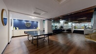 This expansive single-floor recreation facility offers a comprehensive suite of amenities across a generously sized footprint. At its centerpiece is a large indoor swimming pool with vivid turquoise water, framed by distinctive white-tiled support columns and a striking overhead ceiling of warm wood slat beams set against a deep blue-painted structure. Adjacent to the pool is a built-in hot tub, providing a dedicated space for relaxation. A poolside lounge area features vibrant yellow chairs and lounge seating arranged on patterned area rugs, creating a lively and welcoming atmosphere. The fitness center, located off the main pool area, is equipped with a variety of cardio machines and strength-training equipment, including treadmills, ellipticals, and a multi-station cable machine, all set on hardwood-style flooring beneath a vaulted ceiling. A recreation zone with a full-size ping-pong table is accented by a bold navy blue subway tile feature wall with a round mirror. Locker room and restroom facilities are finished with colorful mosaic and square ceramic tile in blue, red, and cream tones, providing a clean and functional space. Throughout the facility, durable flooring and practical lighting ensure a well-maintained, amenity-rich environment.